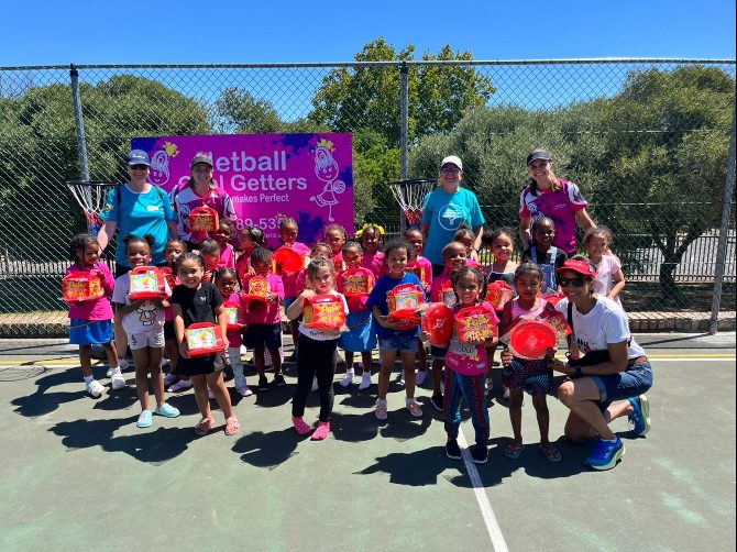 Children holding netball equipment with coaches in front of a Netball Goal Getters banner